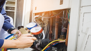 Close-up of person's hands holding manifold gauge set beside exposed AC system components.